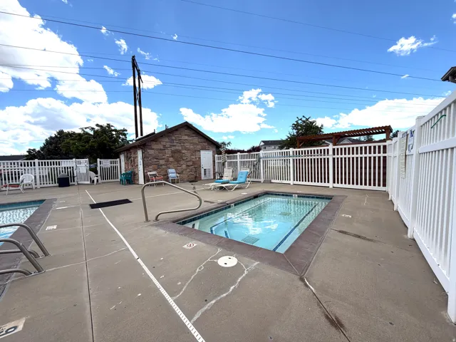 Concrete pool deck featuring two rectangular pools, white picket fencing, and a stone-clad utility building