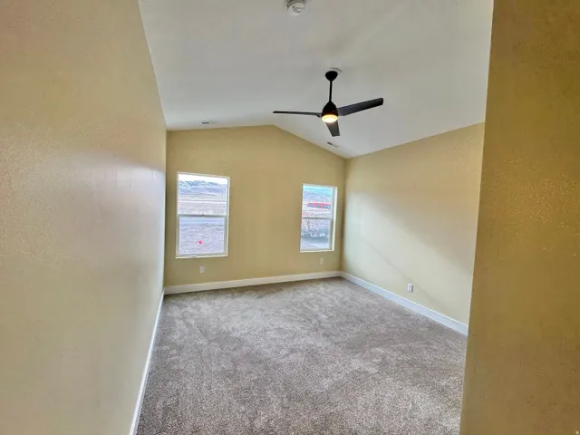 Spacious room featuring vaulted ceilings, a modern ceiling fan with integrated lighting, two windows with white trim, tan painted walls, and grey carpet flooring