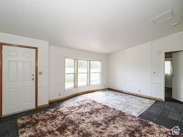 Spacious interior featuring a white four-panel entry door with wood trim, multiple windows providing natural light, and contrasting dark flooring