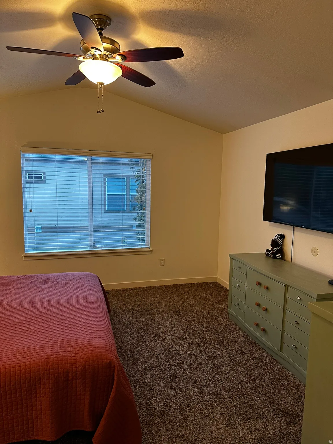 Vaulted ceiling room featuring a ceiling fan with integrated lighting, a large window with blinds, and neutral-toned wall-to-wall carpeting