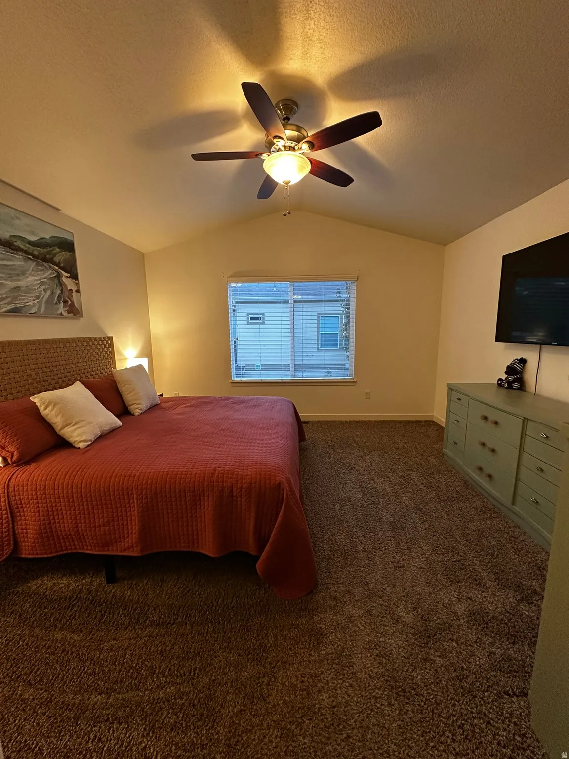 Carpeted interior space featuring a vaulted ceiling with a ceiling fan light fixture, a window with blinds, and a built-in wall-mounted television