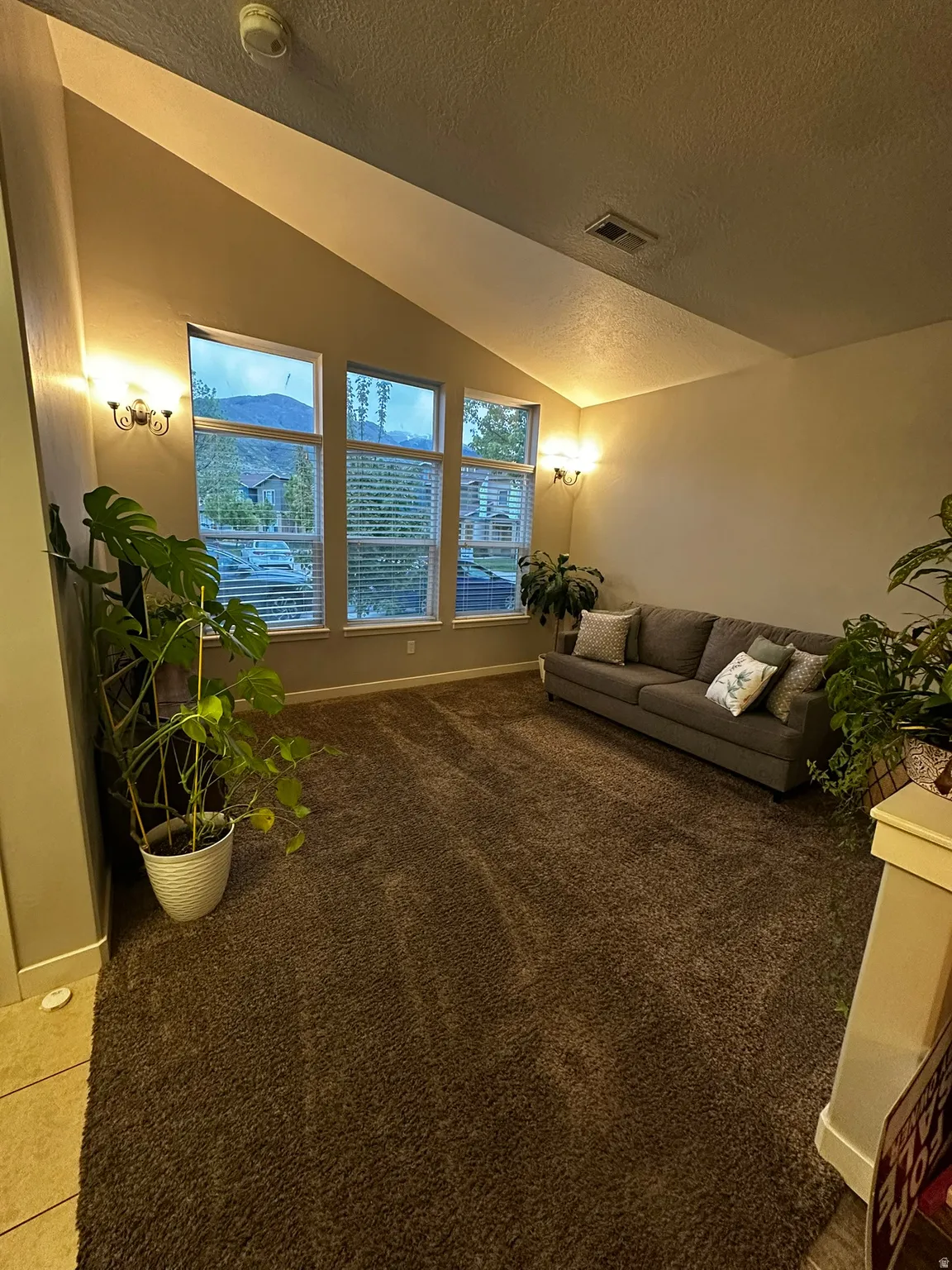 Vaulted ceiling living area featuring a trio of large windows with blinds, sconce lighting, and brown carpeting