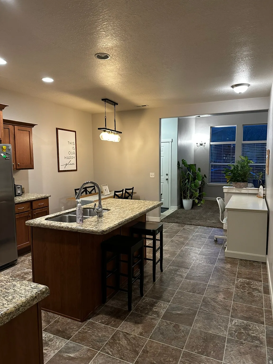 Kitchen featuring a central island with an integrated sink, granite-finish countertops, wood-finish cabinetry, and square tile flooring