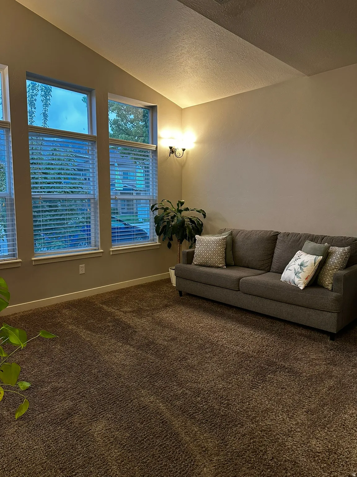 Vaulted ceilings accent a living space featuring multiple windows with blinds, a wall-mounted sconce light fixture, and textured carpet flooring