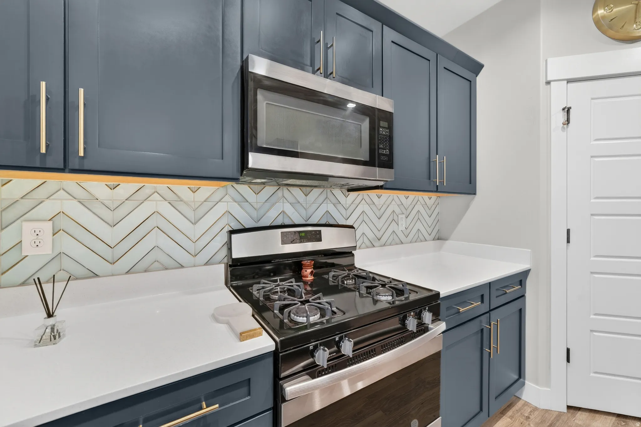 Modern kitchen featuring dark blue cabinetry with gold-tone hardware, stainless steel appliances, white countertops, and a chevron tile backsplash