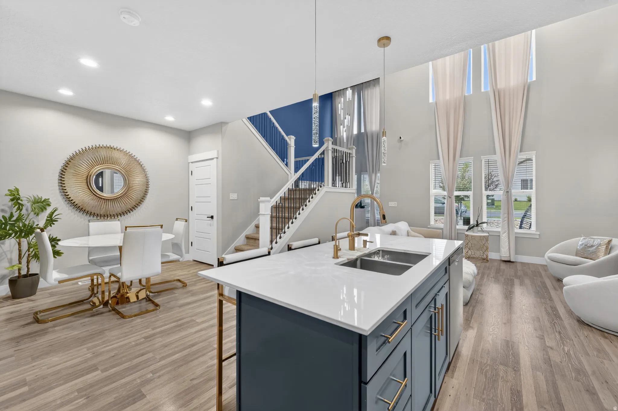 Open-concept living area featuring a kitchen island with dual basin sink and gold-tone faucet, wood-finish flooring, and a dramatic two-story ceiling with tall windows