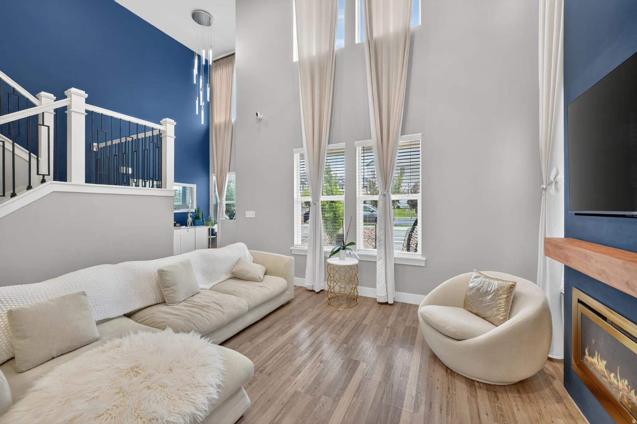 Spacious living area featuring high ceilings, tall windows with blinds, wood-finish flooring, a contemporary fireplace with a wood mantel, and a staircase with white balusters and black metal spindles