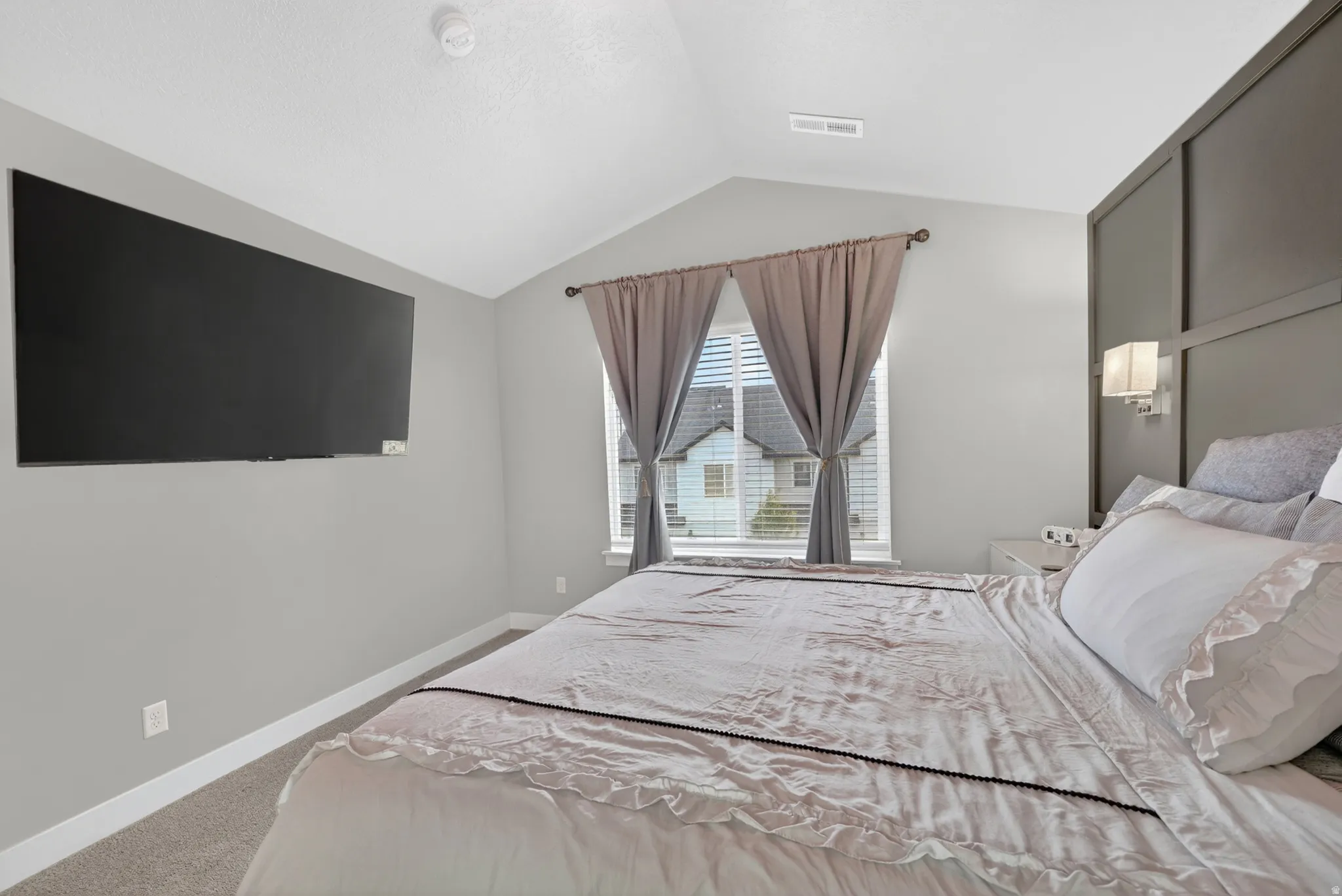 Vaulted ceiling bedroom with light gray walls, plush carpet, and a window featuring blinds and drapes