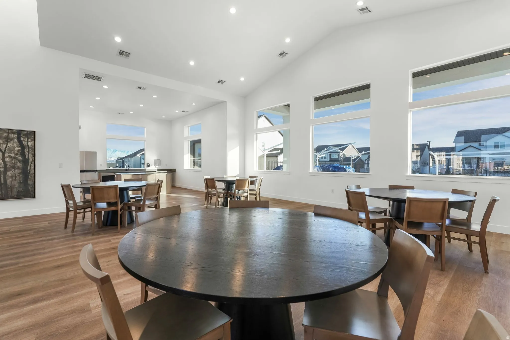 Spacious common area featuring vaulted ceilings, abundant natural light from large windows, wood-finish flooring, recessed lighting, and multiple round tables with seating