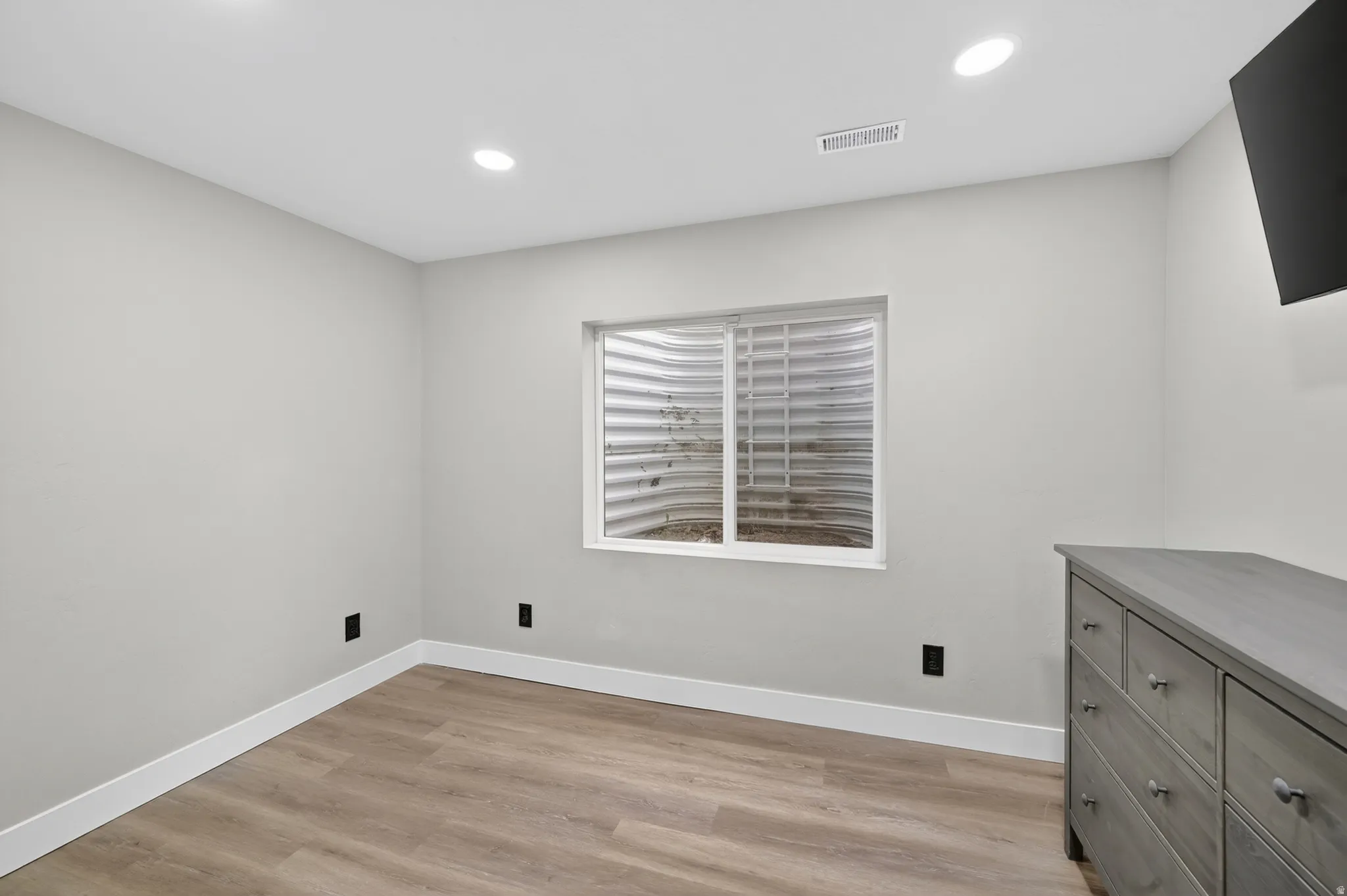Room featuring light gray walls, wood-finish flooring, recessed ceiling lighting, and a white-framed egress window with a corrugated metal window well