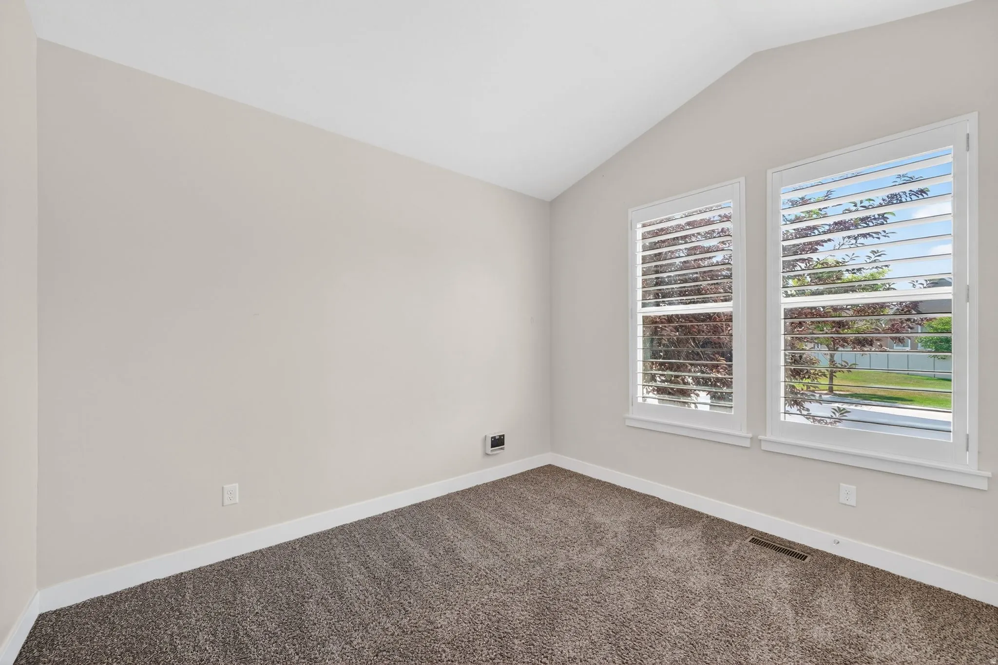 Vaulted ceiling room featuring two windows with white plantation shutters, neutral-toned walls, dark gray carpet, and white baseboards