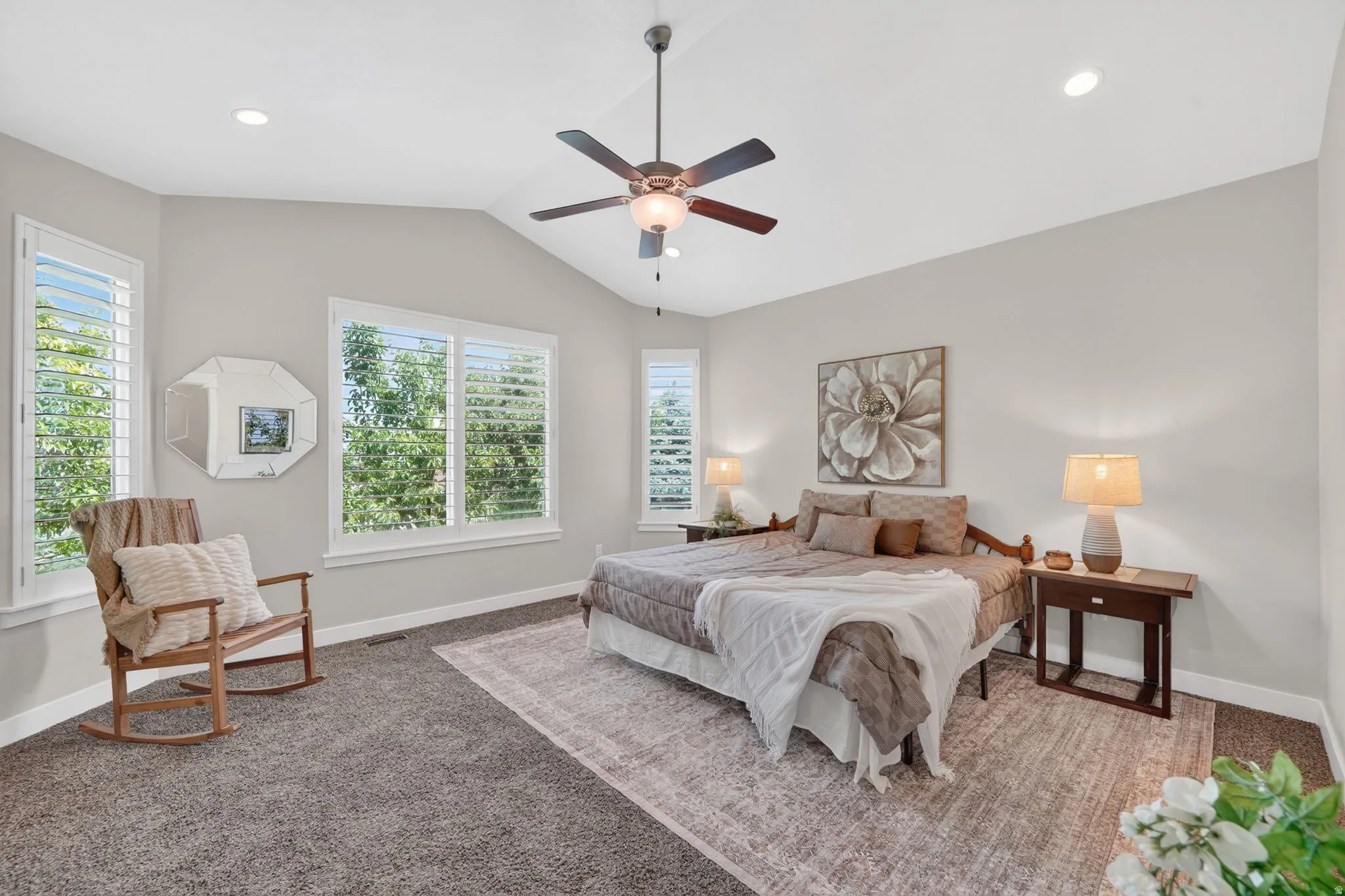 Spacious bedroom featuring vaulted ceilings, recessed lighting, a ceiling fan, multiple windows with plantation shutters, and dark gray carpeting
