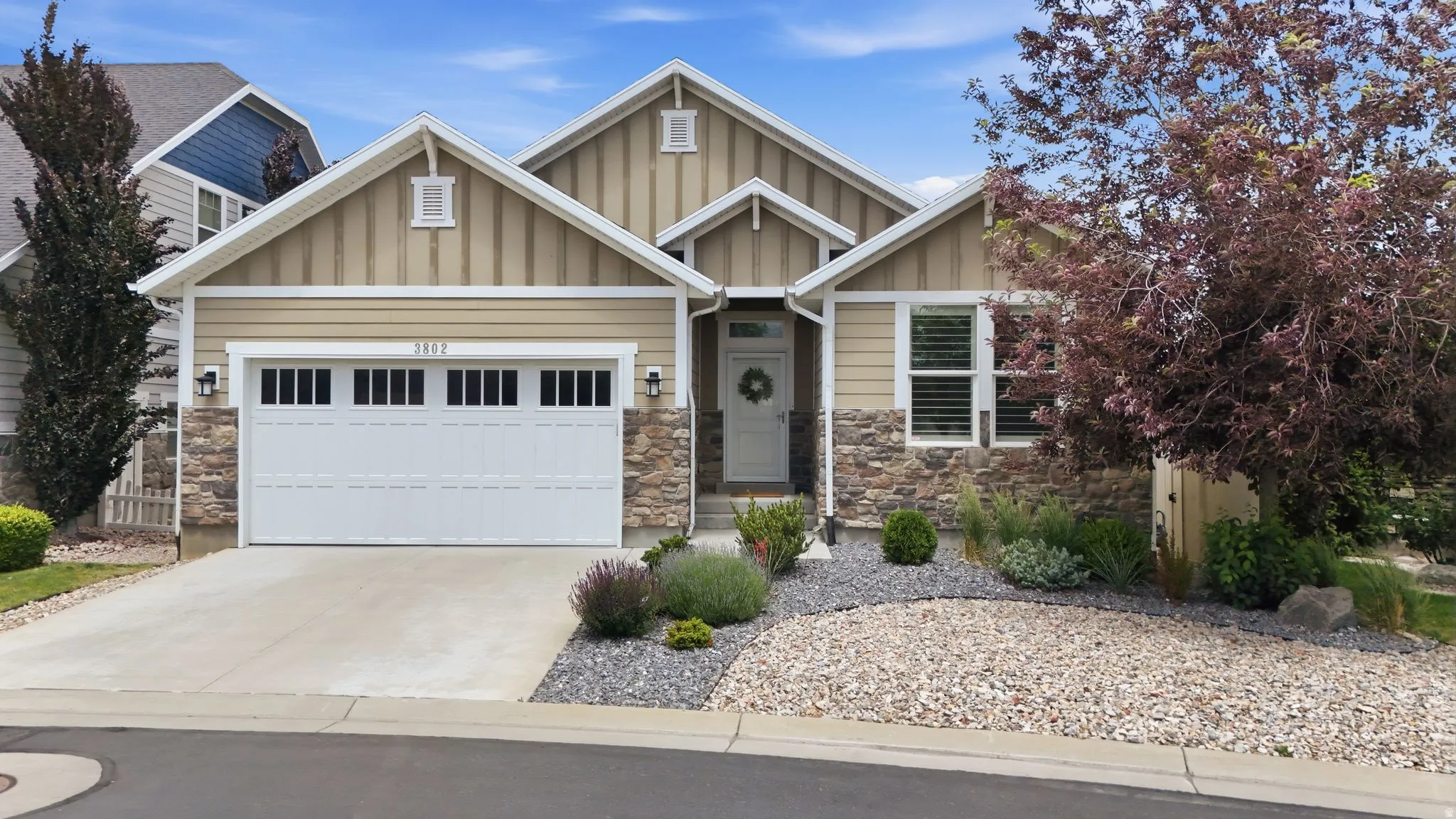 Inviting home exterior featuring a two-car garage with transom windows, board and batten siding, stone facade accents, a concrete driveway, and a low-maintenance gravel and shrub landscape