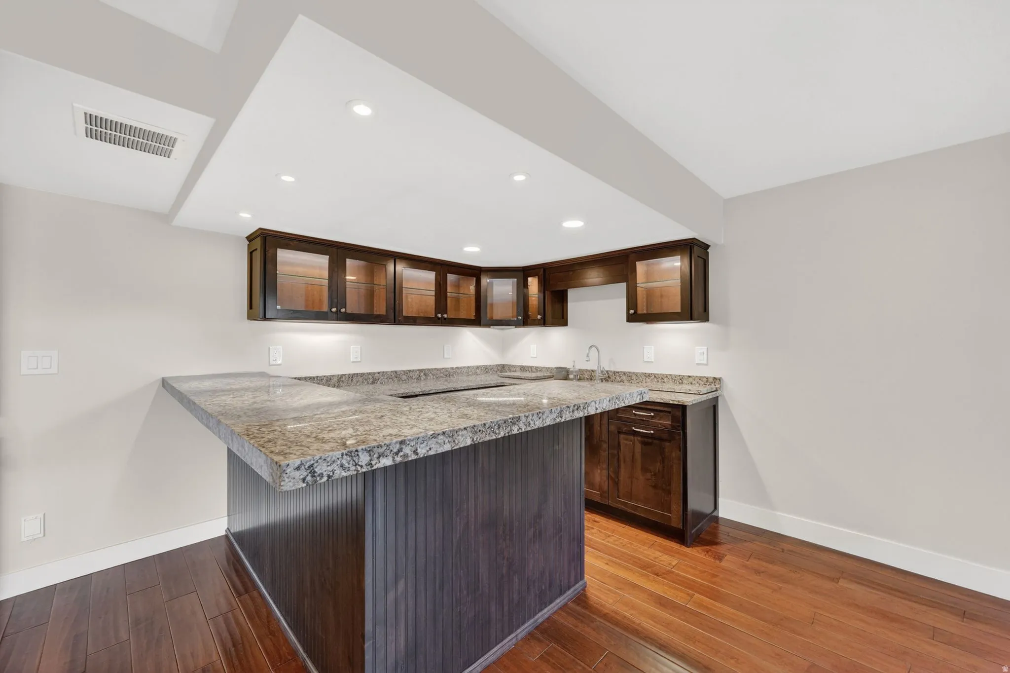 Wet bar featuring wood-finish flooring, dark wood cabinetry, granite countertops, and recessed lighting