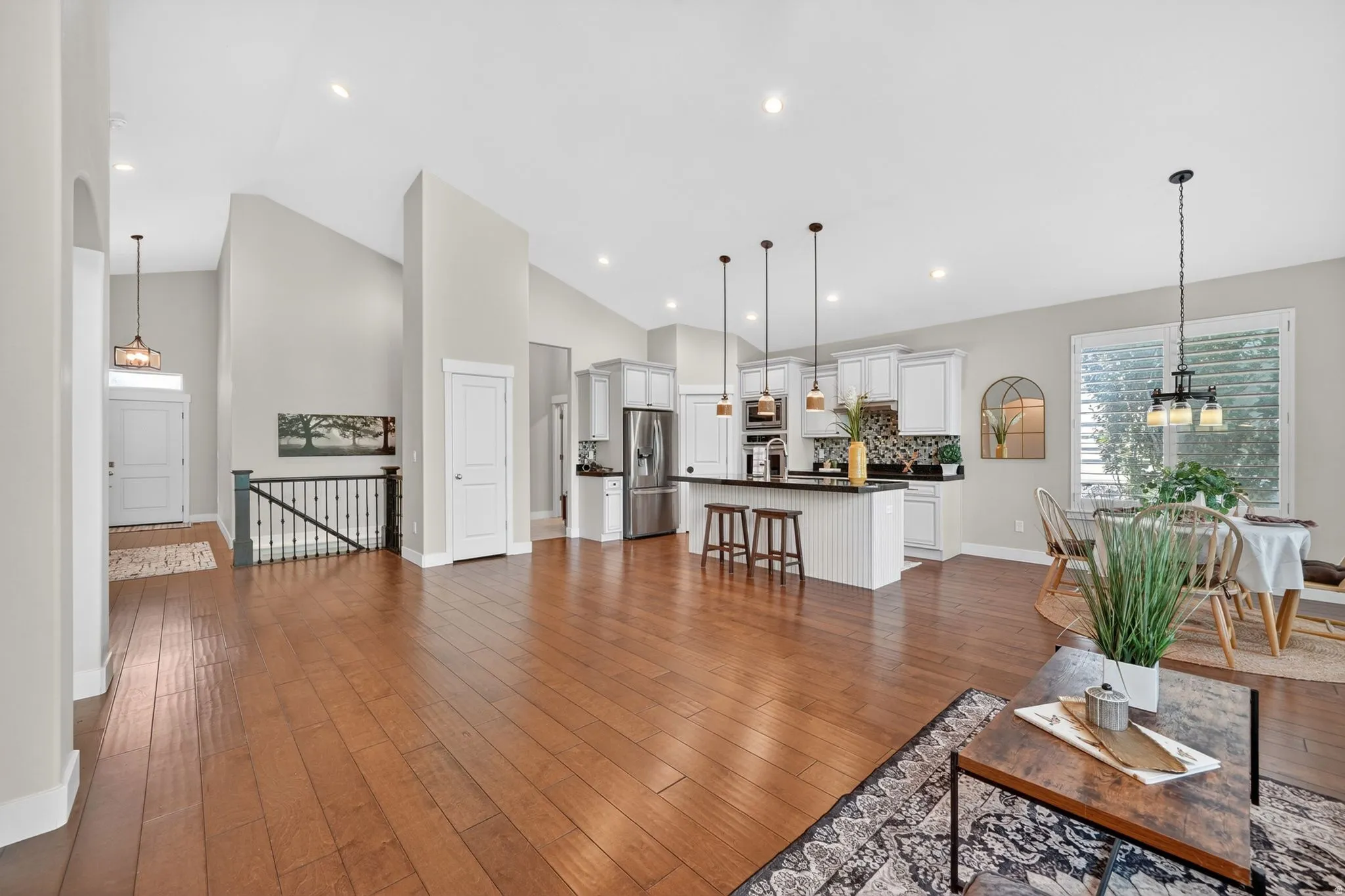 Open-concept living space featuring hardwood flooring, vaulted ceilings, a kitchen with white cabinetry and stainless steel appliances, a kitchen island with pendant lighting, and a dining area with shuttered windows