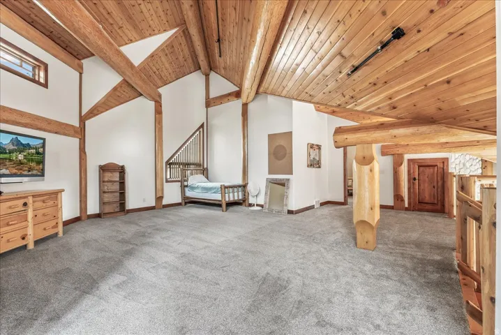 Spacious room featuring exposed wood beams, vaulted wood plank ceilings, plush gray carpeting, white walls, and a built-in wood railing