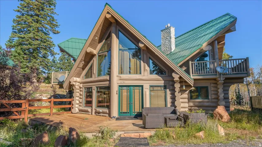 Log cabin exterior featuring a prominent A-frame design, a green metal roof, a stone chimney, extensive window-paneled walls, and a wooden deck