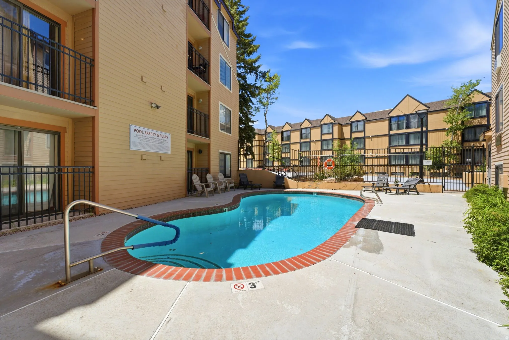 Outdoor swimming pool with concrete decking and brick coping, surrounded by multi-story residential buildings with balconies and a fenced perimeter