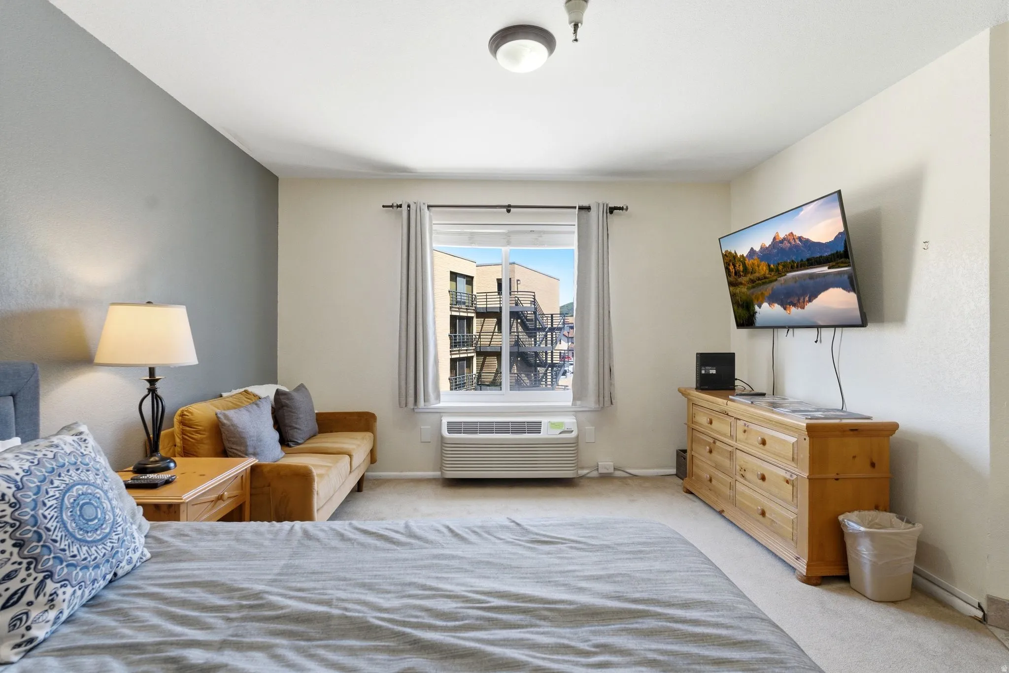 Carpeted room with a feature wall, window with exterior building visibility, and a wall-mounted climate control unit