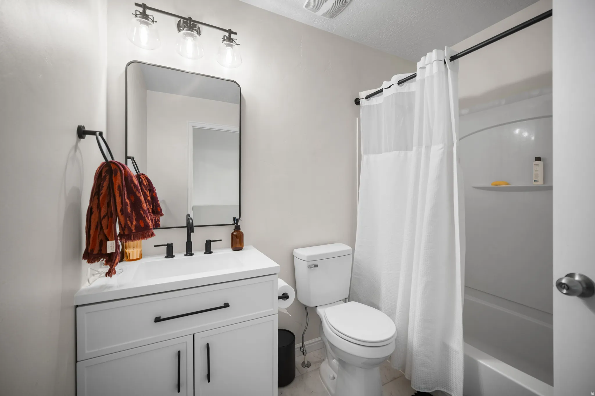 Bathroom featuring a white vanity with black hardware, a rectangular mirror, a three-light fixture, and a white tub with shower