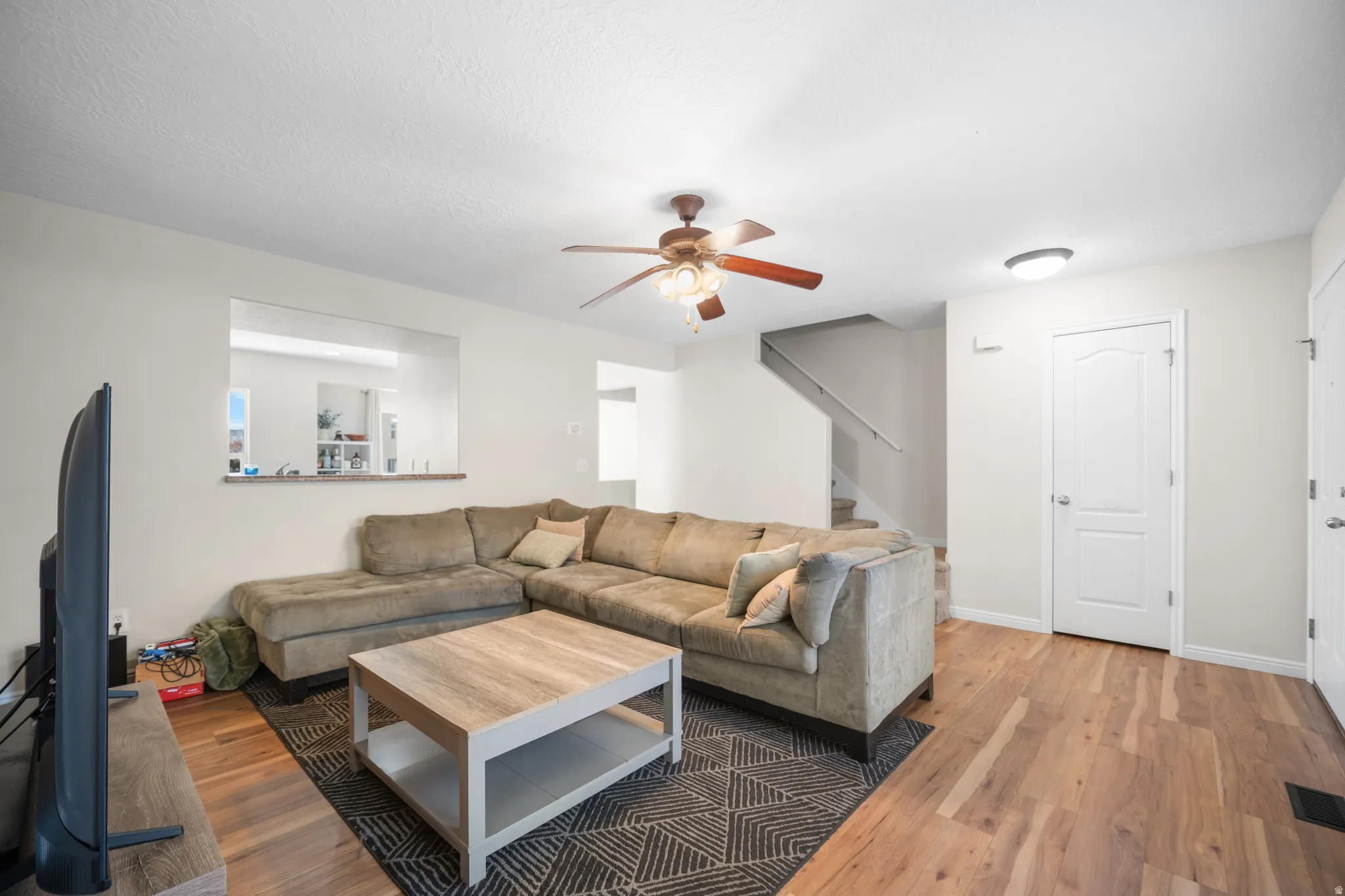 Living area featuring light wood-finish flooring, a ceiling fan with integrated lighting, and a pass-through window to an additional space