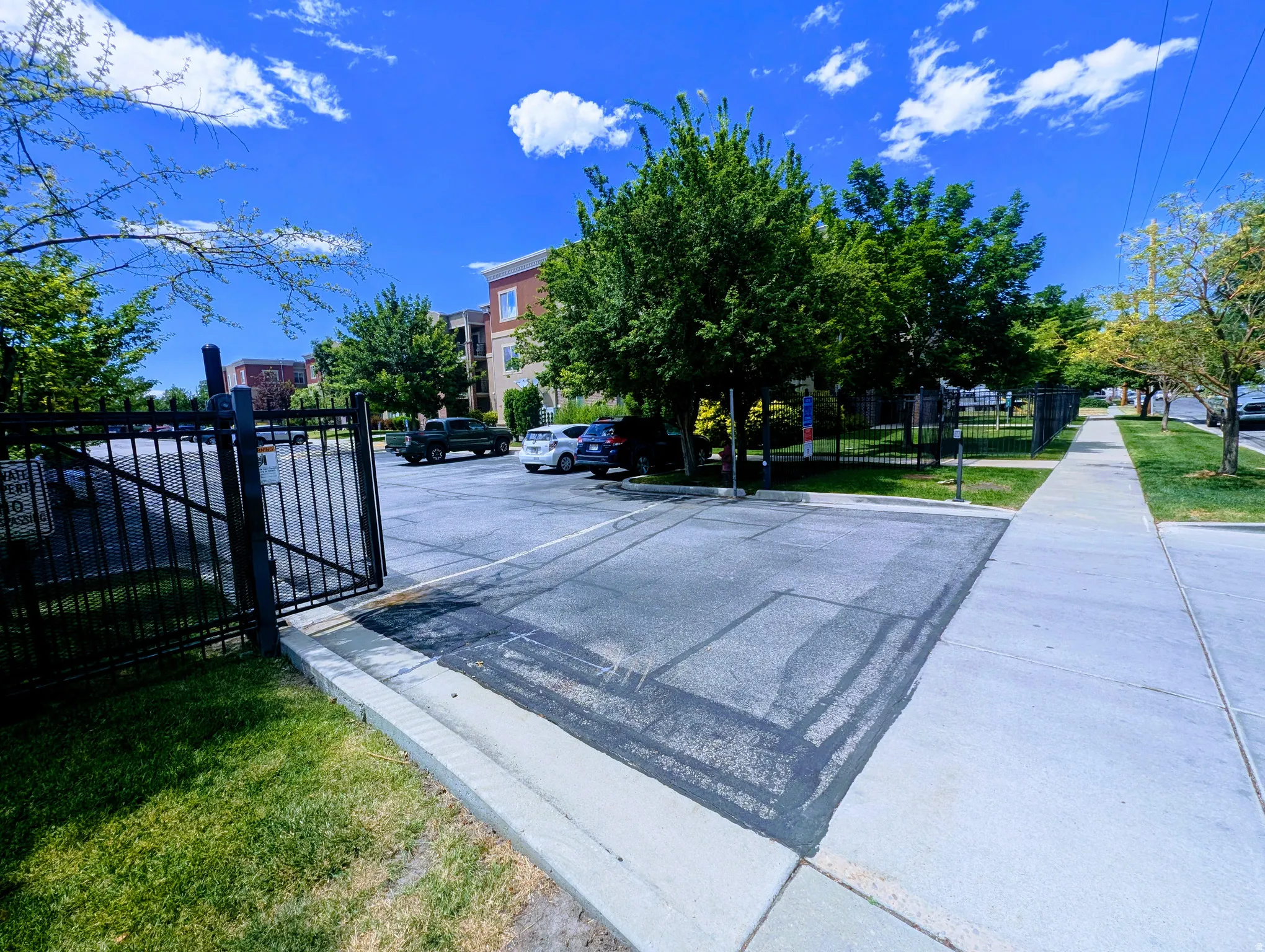 Paved parking area featuring a black metal gate and fence, surrounded by mature trees and a manicured lawn