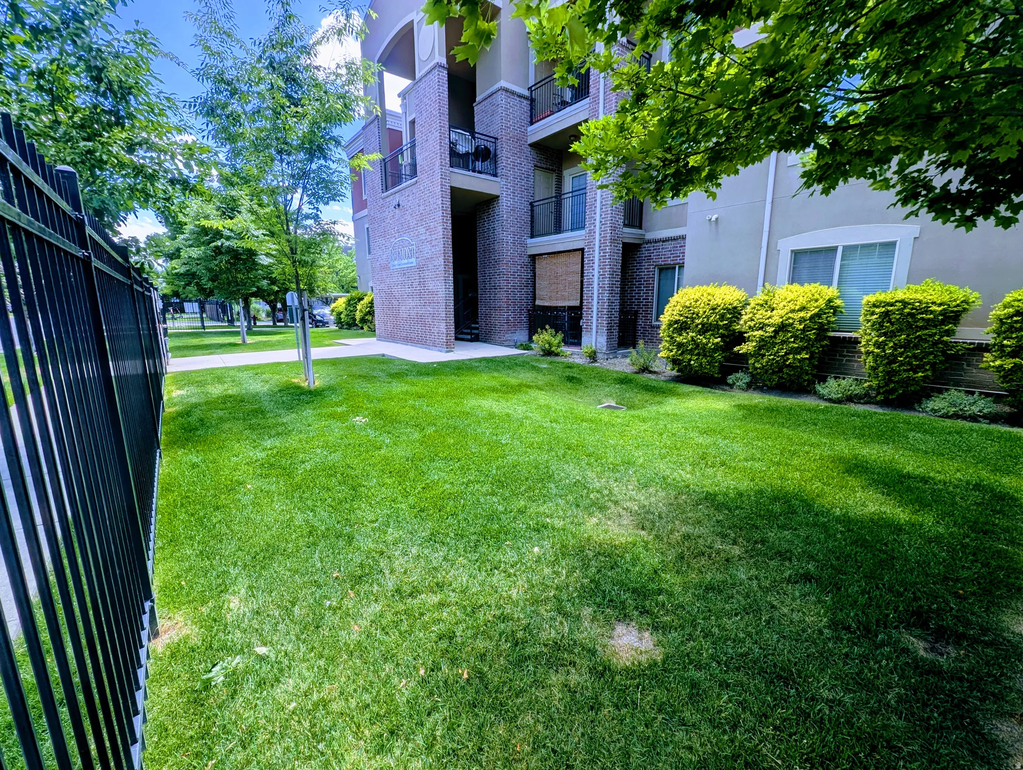 Green lawn area with a black metal fence, bordered by a multi-story building featuring brick accents and private balconies