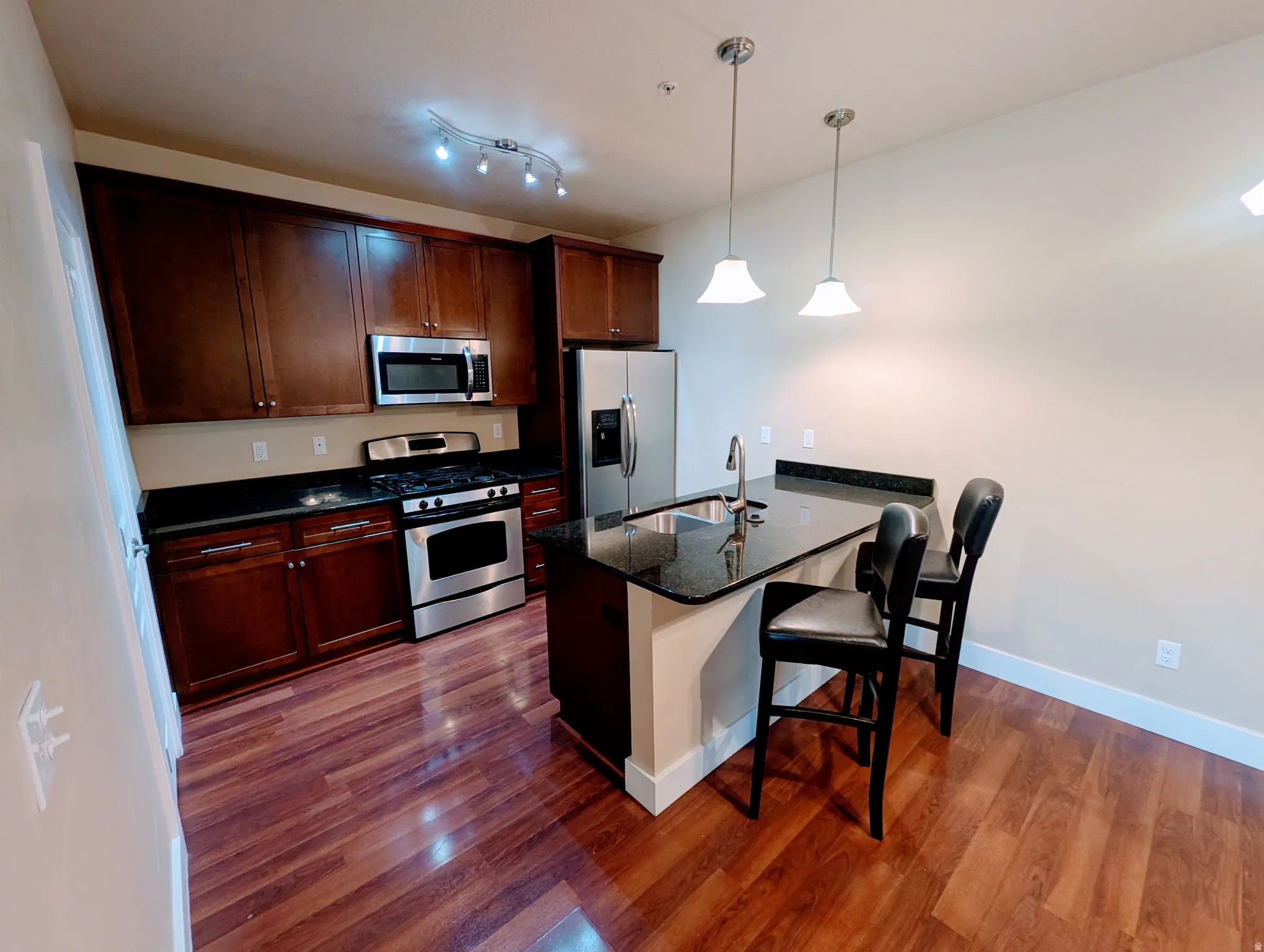 Kitchen featuring rich wood-finish cabinetry, stainless steel appliances, dark stone countertops, and a peninsula with pendant lighting
