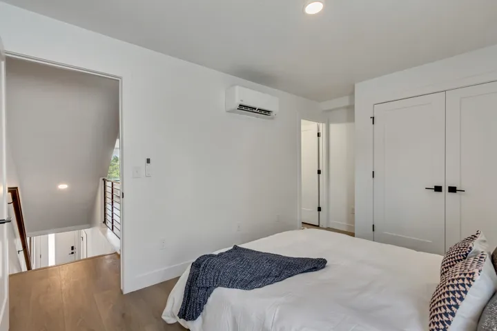 Contemporary bedroom featuring a white interior palette, wood-finish flooring, two panel doors with matte black hardware, recessed lighting, and a wall-mounted mini-split HVAC unit