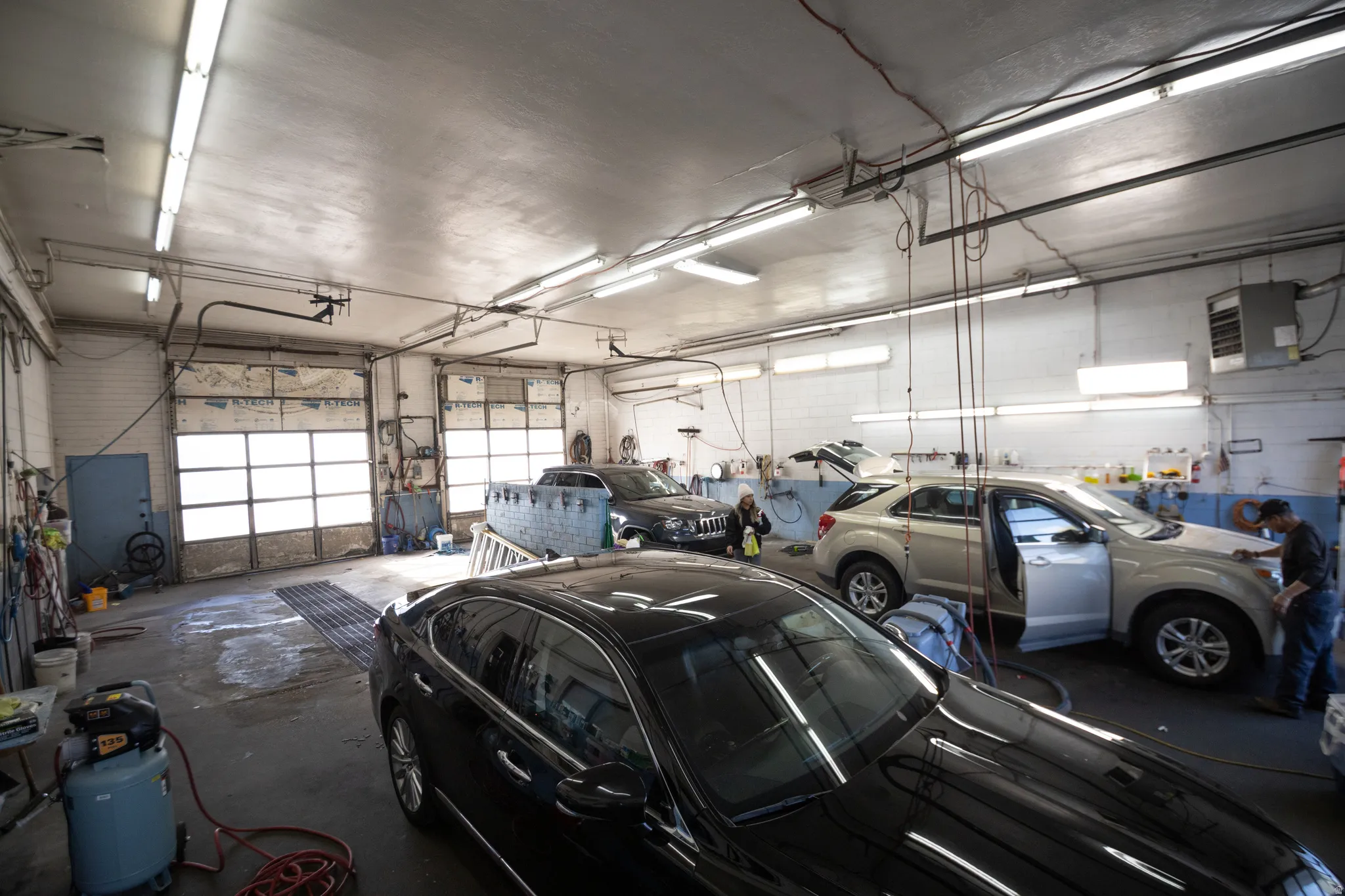 Spacious commercial garage featuring two overhead bay doors, concrete flooring, exposed overhead lighting, a wall-mounted heating unit, and a side access door