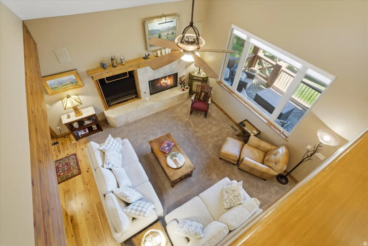 Aerial view of spacious living area featuring a stone-clad fireplace with built-in shelving, a natural wood mantle, a ceiling fan, and large windows with transom panels