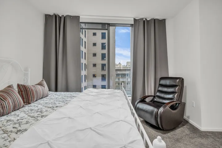 Room featuring a large window with city buildings visible, neutral-toned walls, and grey floor-covering