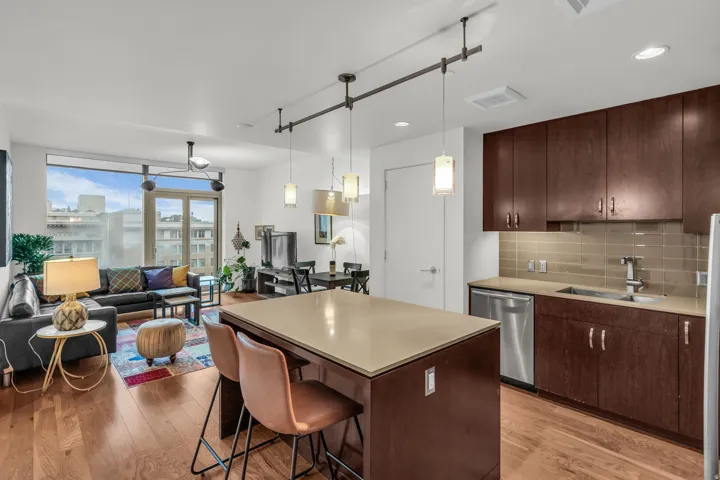 Open-concept living area featuring wood-finish flooring, a kitchen island with a light-toned countertop, dark wood-finish cabinetry, stainless steel appliances, and a tiled backsplash