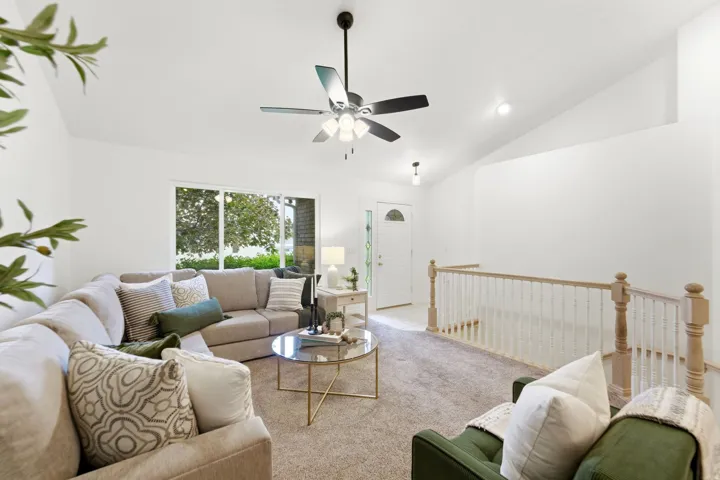 Bright living area featuring vaulted ceilings, a ceiling fan, plush carpeting, and a window overlooking exterior greenery