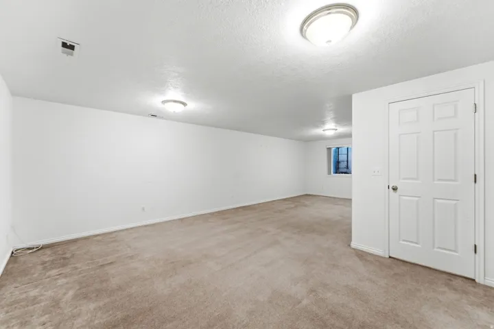 Spacious room featuring neutral carpet flooring, white walls, flush-mount ceiling lighting, a four-panel door, and a window with a security grate