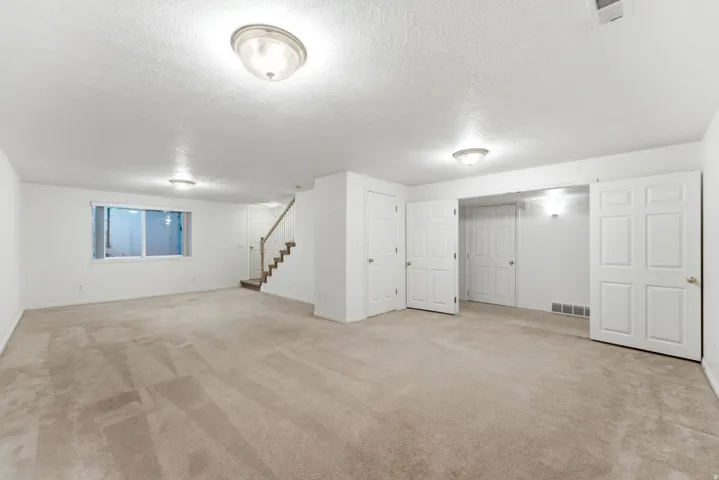 Spacious room featuring neutral carpet, white walls, multiple flush-mount ceiling lights, a large window, and a staircase with wood-finish handrails