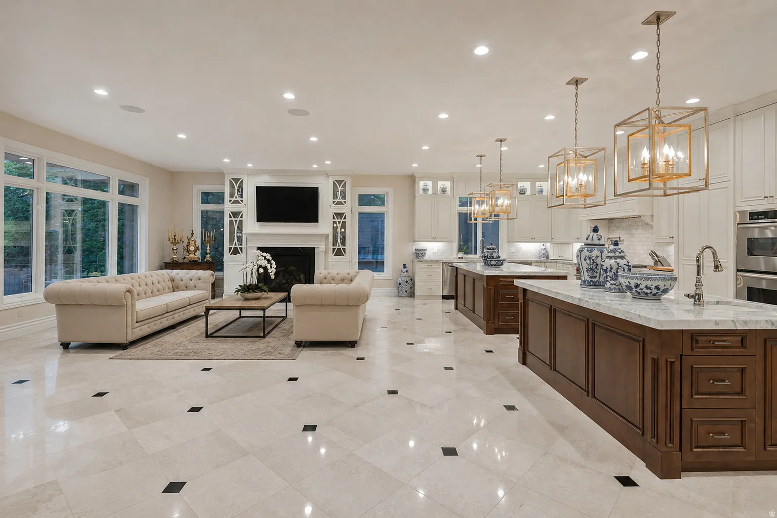 Expansive open-concept living space featuring light-toned tile flooring with dark accent squares, a fireplace with a white mantel and built-in shelving, large windows, two kitchen islands with dark wood cabinetry and light countertops, and multiple pendan