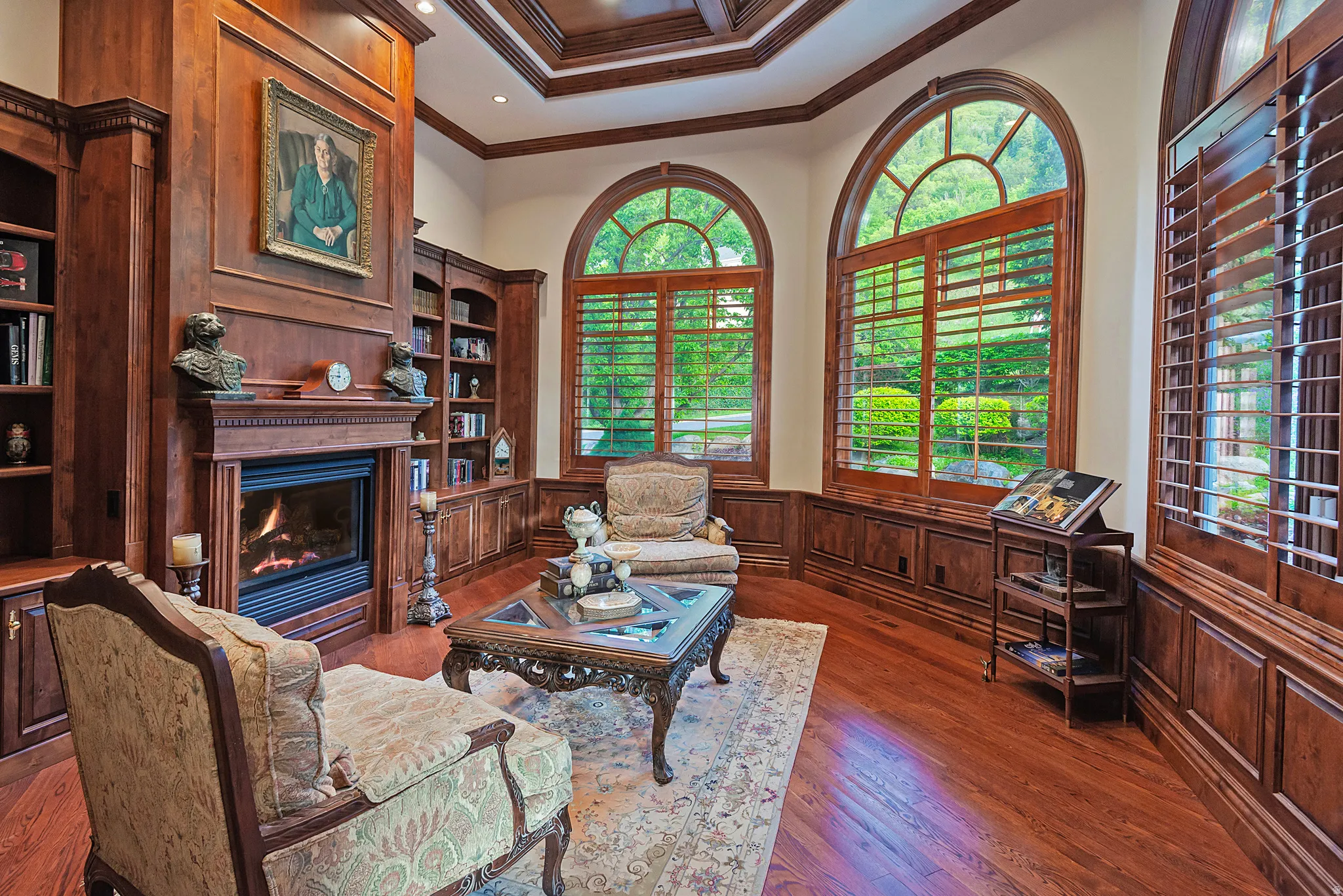 Rich wood-paneled walls featuring built-in shelving, a gas fireplace with a decorative mantel, and arched windows with plantation shutters
