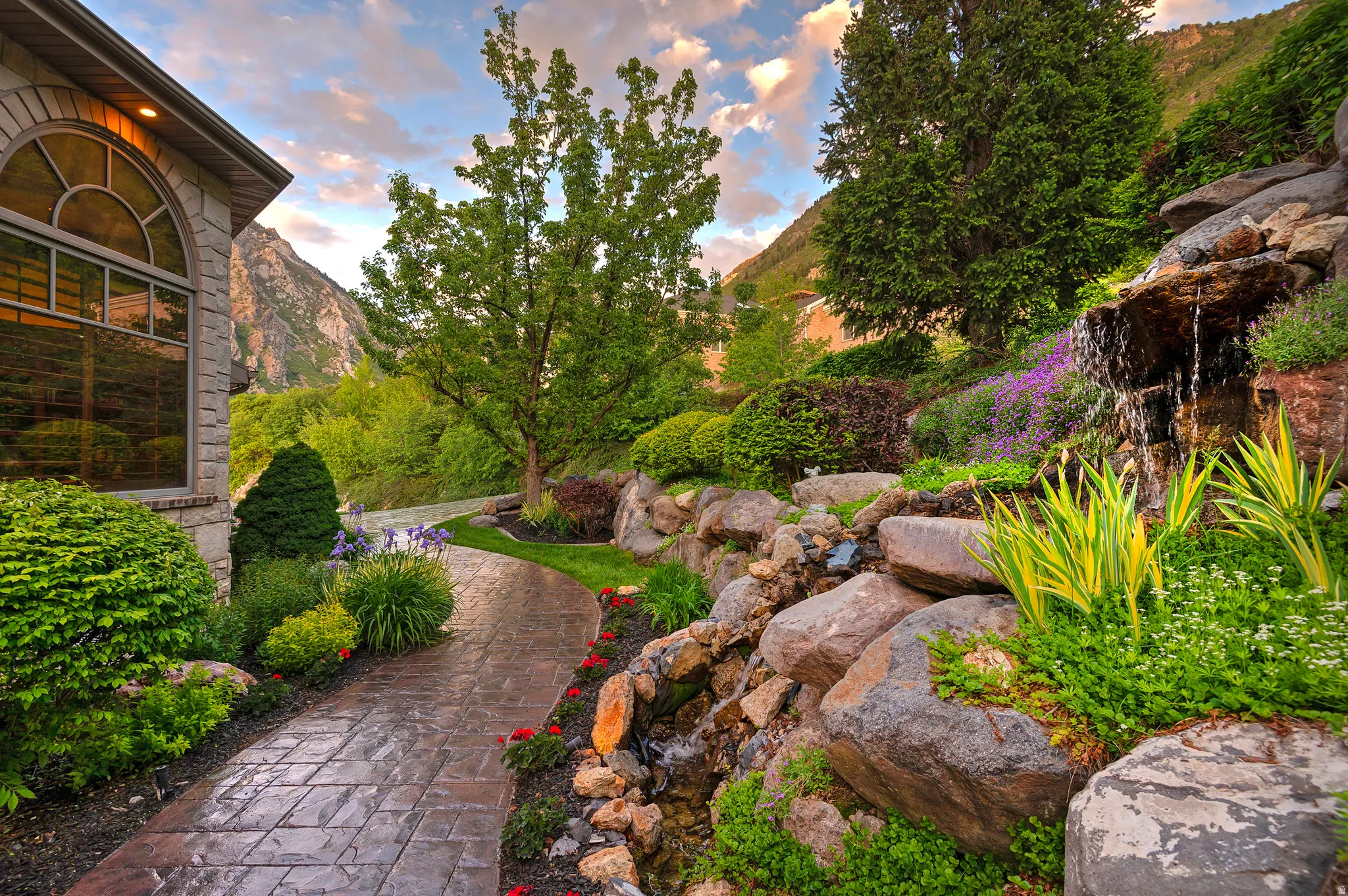 Professionally landscaped grounds featuring a stone waterfall, mature trees, and a stamped concrete pathway
