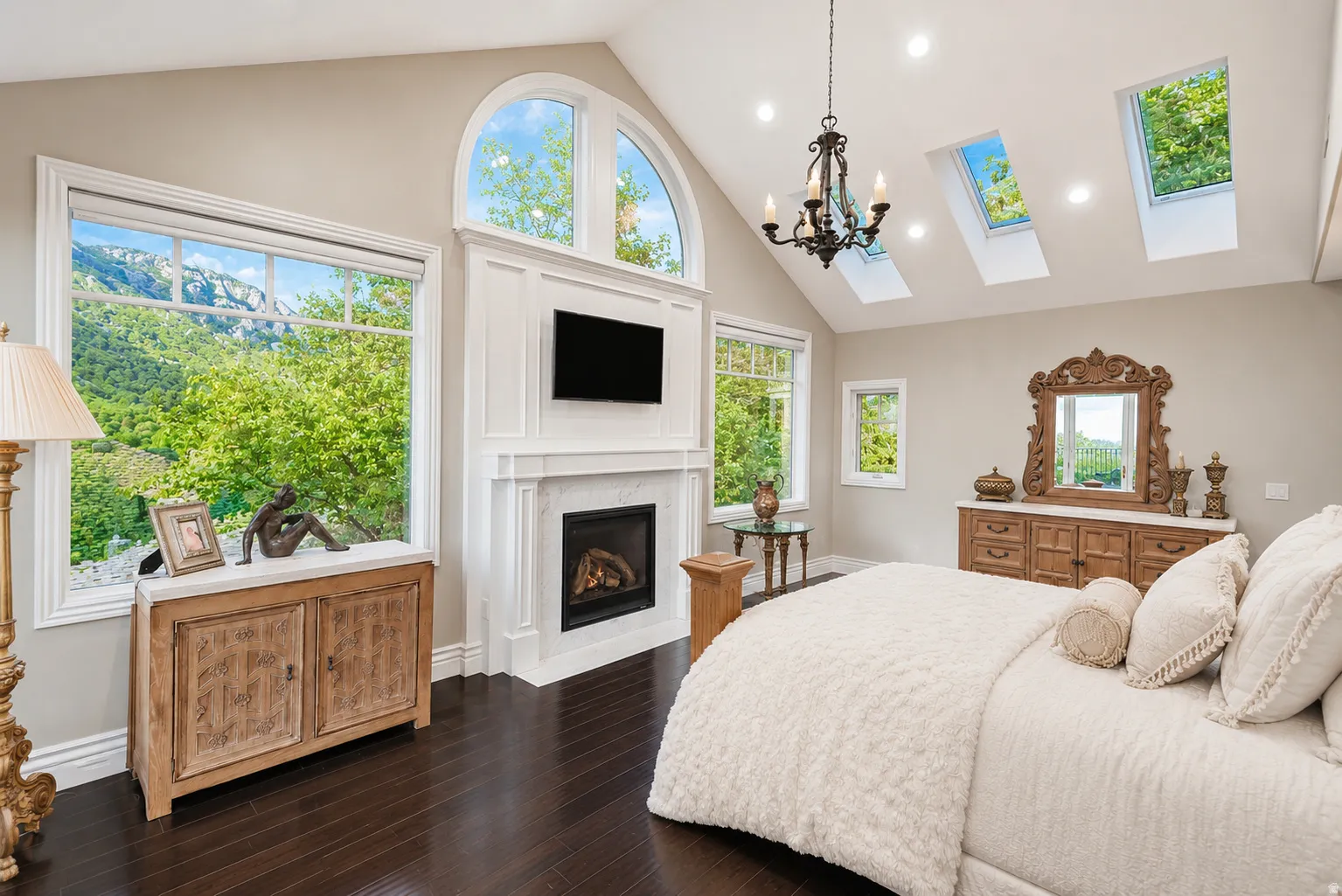 Vaulted ceiling bedroom with dark wood-finish flooring, a white fireplace with a wall-mounted screen, and abundant natural light from multiple windows and skylights