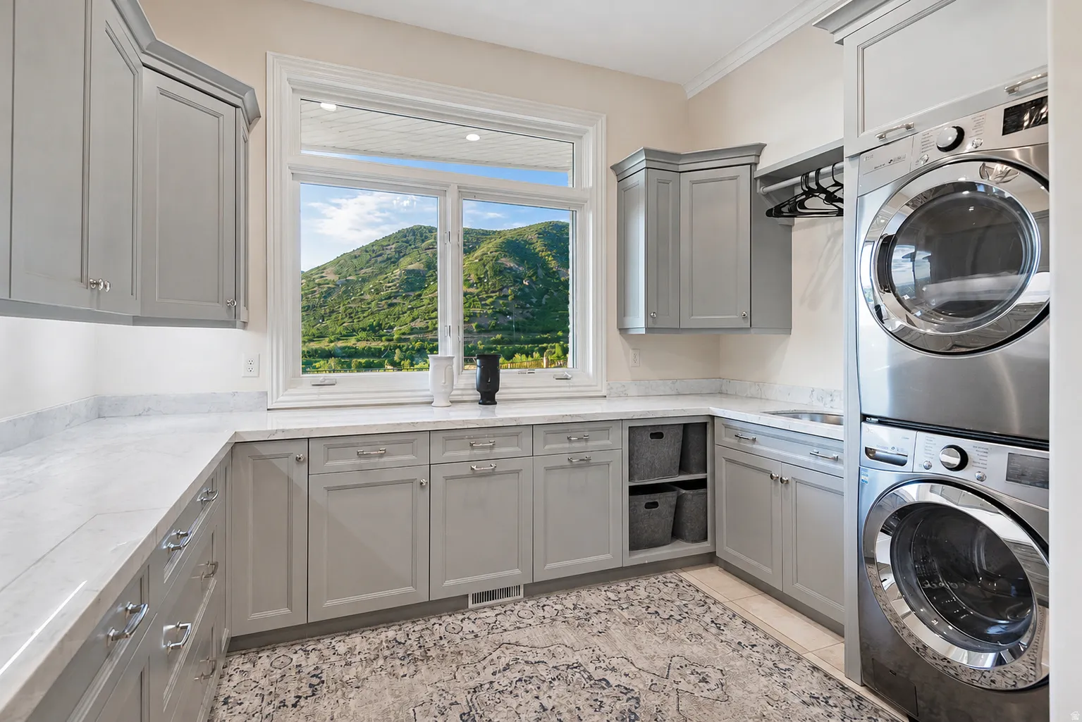 Bright utility room featuring ample light gray cabinetry, white countertops, built-in shelving, a stainless steel stacked washer and dryer, and a large window overlooking a verdant hillside