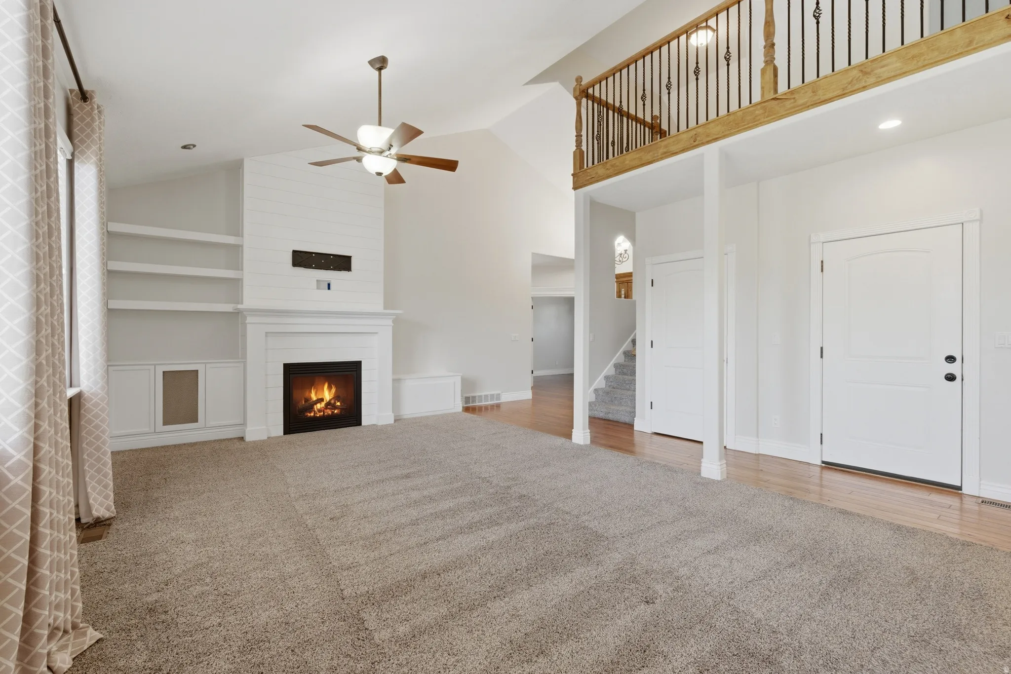 Vaulted living area featuring a fireplace with shiplap surround, built-in shelving, ceiling fan, carpet flooring, and an upper-level wood-railed overlook