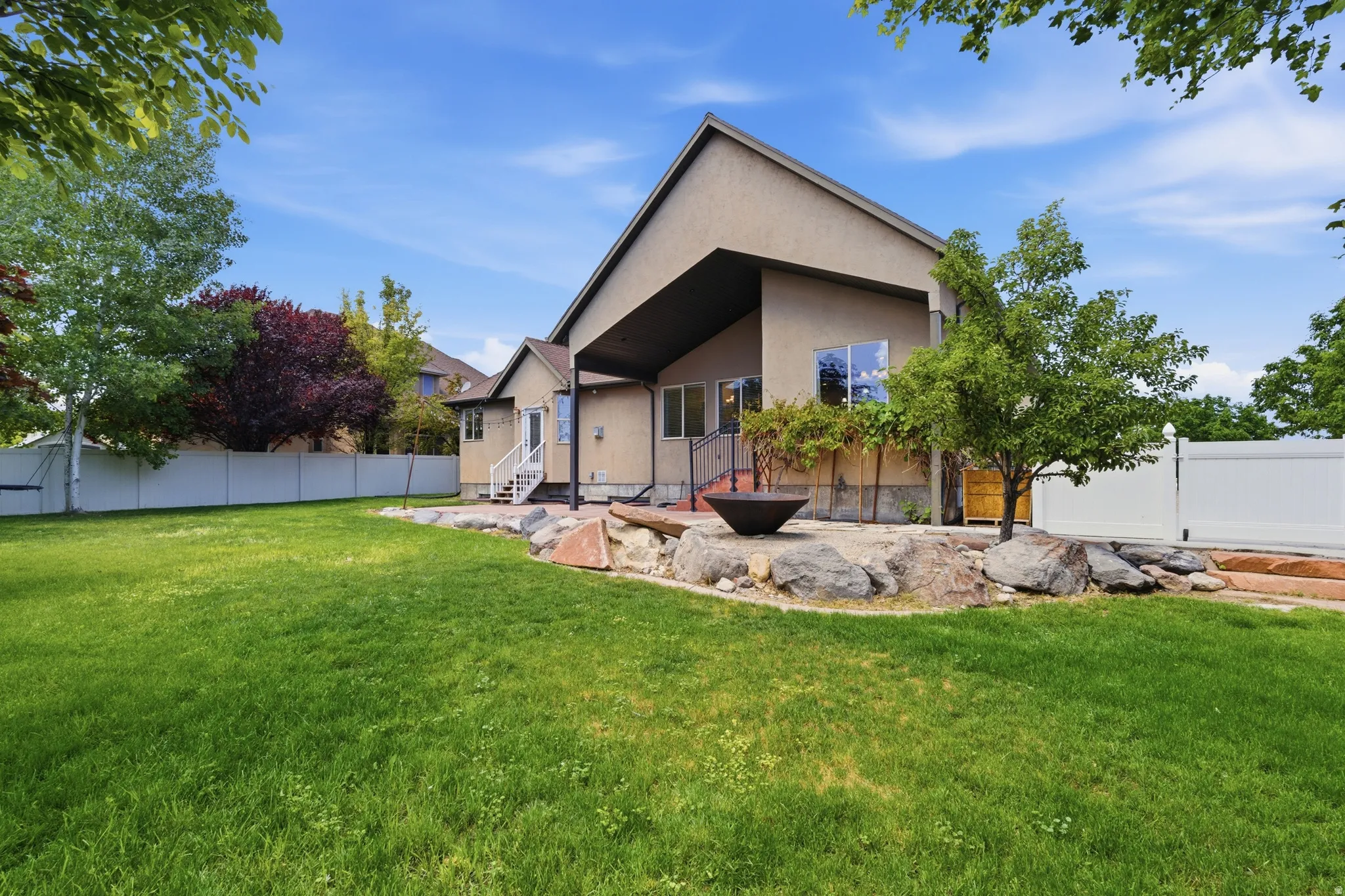 Expansive backyard featuring a manicured lawn, rock landscaping with a fire pit, mature trees, and privacy fencing