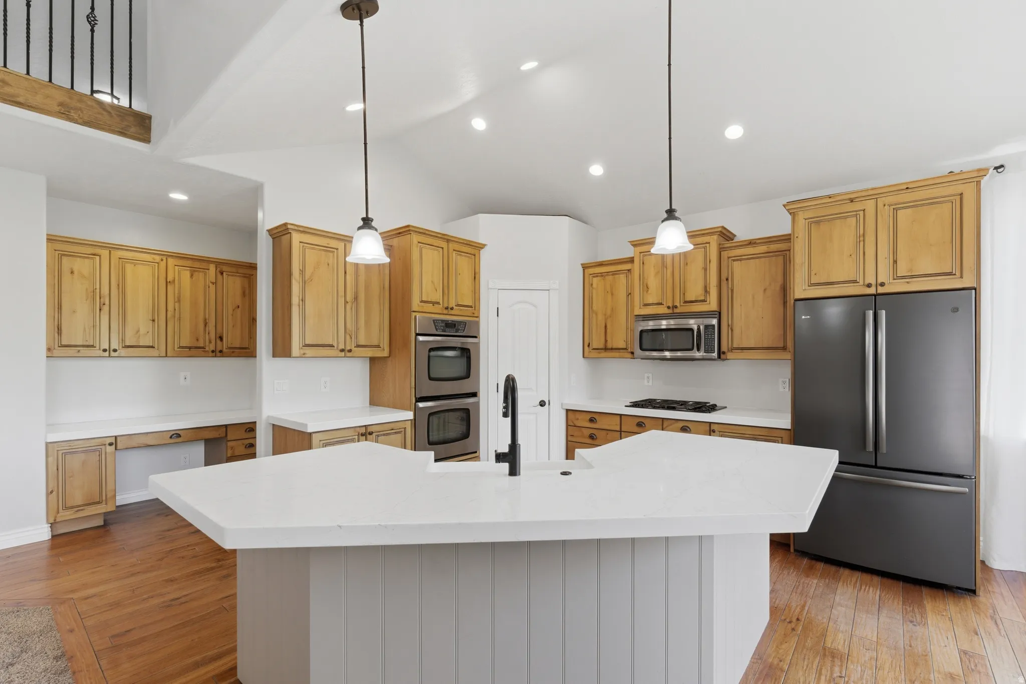 Kitchen featuring a large central island with a white countertop and pendant lighting, wood-finish flooring, light wood cabinetry, double wall ovens, and a dark stainless steel refrigerator