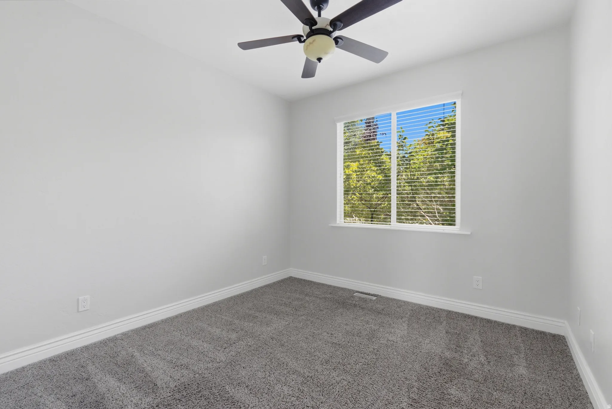 Bright room featuring a ceiling fan with light fixture, neutral gray carpeting, white baseboards, and a window with horizontal blinds offering a green outdoor scenery