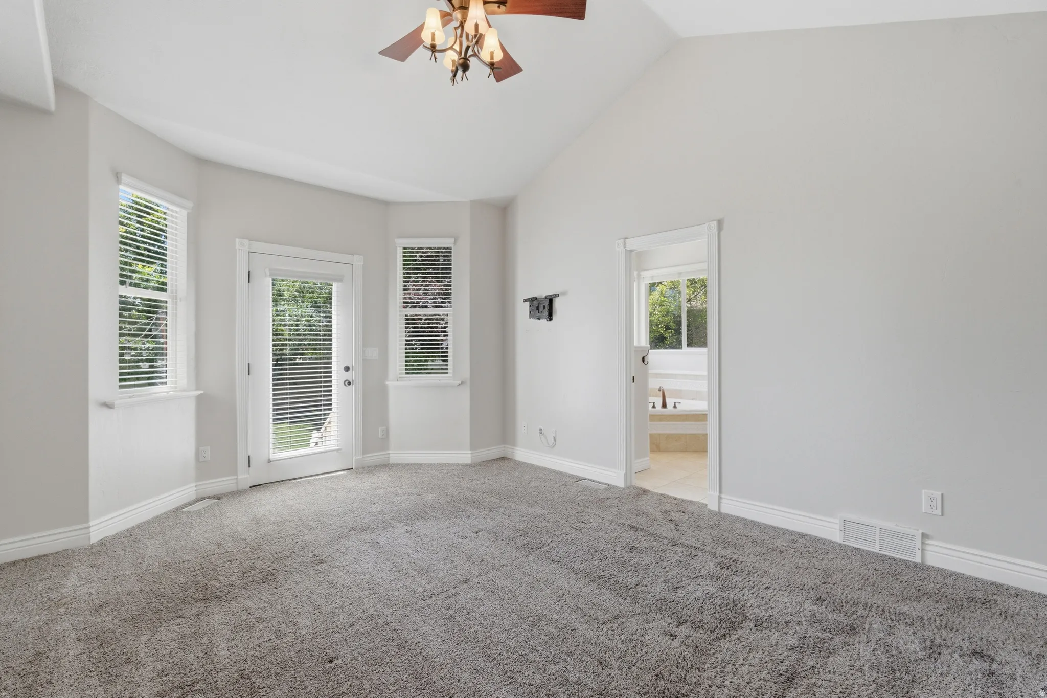 Carpeted room featuring a vaulted ceiling with a ceiling fan, a private outdoor access door, two windows with blinds, and an open doorway revealing a portion of a tiled bathroom with a built-in tub