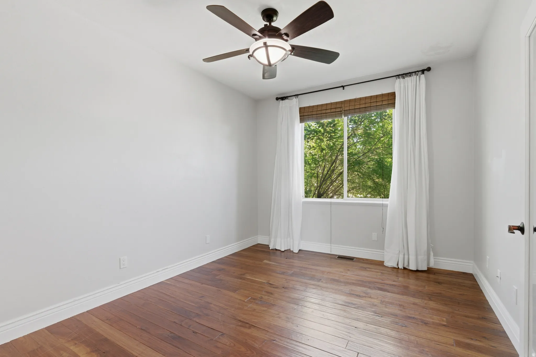 Room with wood-finish flooring, a window with a woven wood blind, and white baseboards