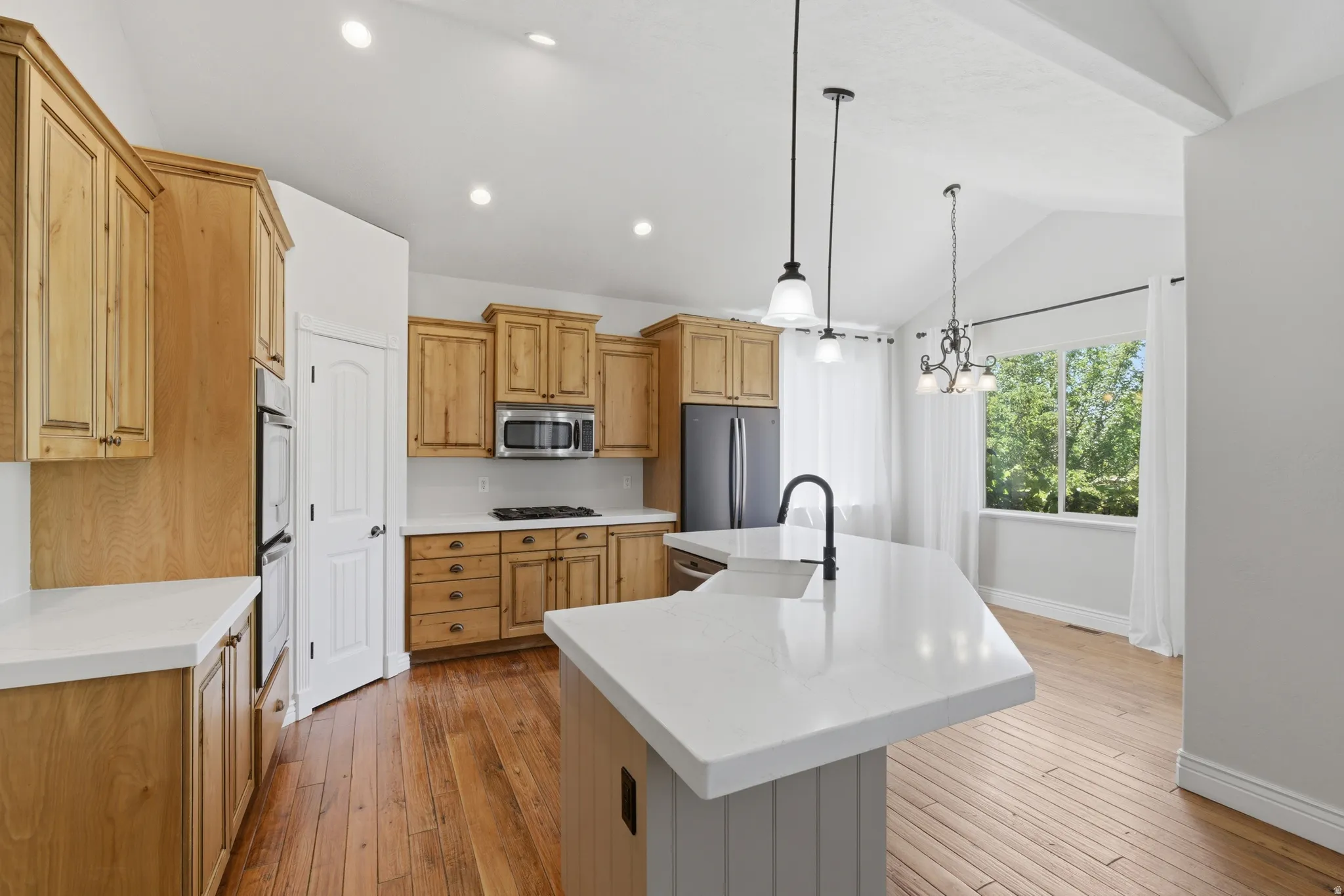 Spacious kitchen featuring light wood cabinetry, white countertops, built-in double ovens, a central island with sink, and wood-finish flooring