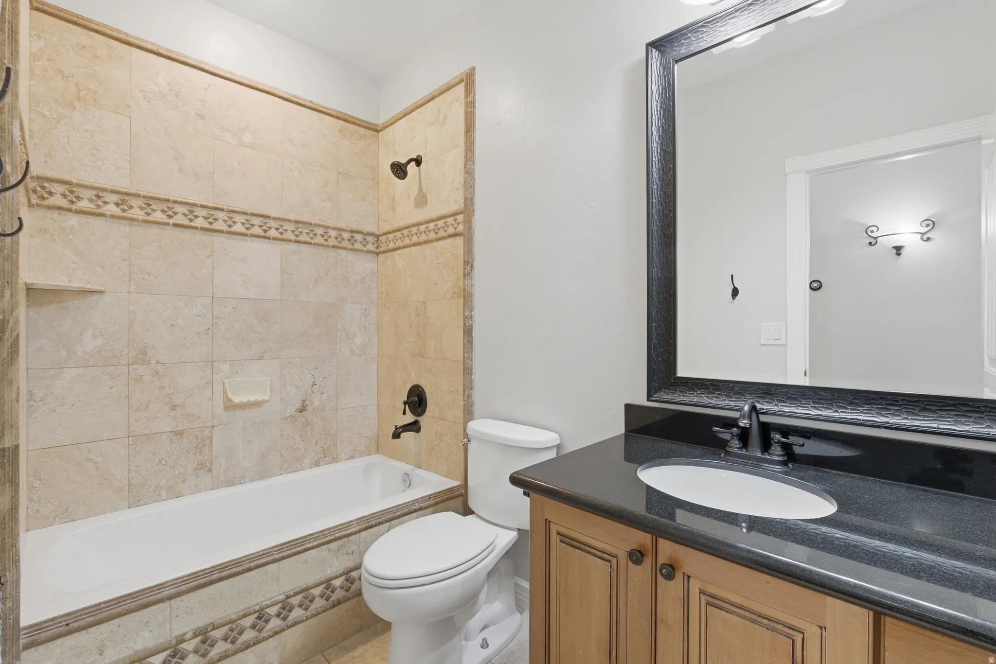 Bathroom featuring a bathtub with tiled surround, a dark stone-finish countertop, wood-finish cabinetry, an oval undermount sink, and a framed mirror