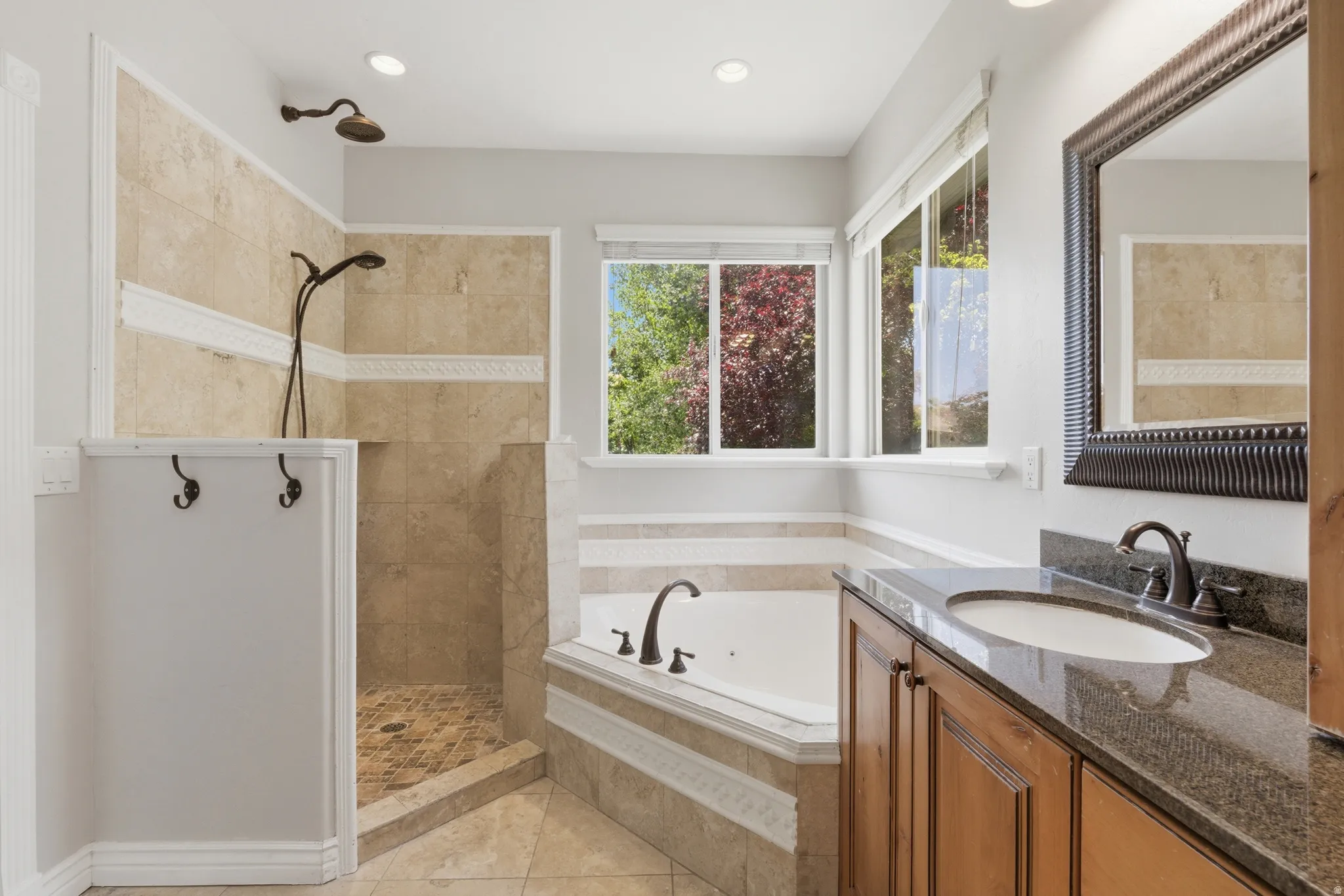 Bathroom featuring a tiled walk-in shower with dual showerheads, a corner soaking tub, a double window, a single vanity with a dark stone countertop, and a framed mirror